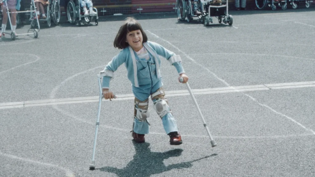 A smiling young girl using crutches and leg braces stands on a playground with other children.