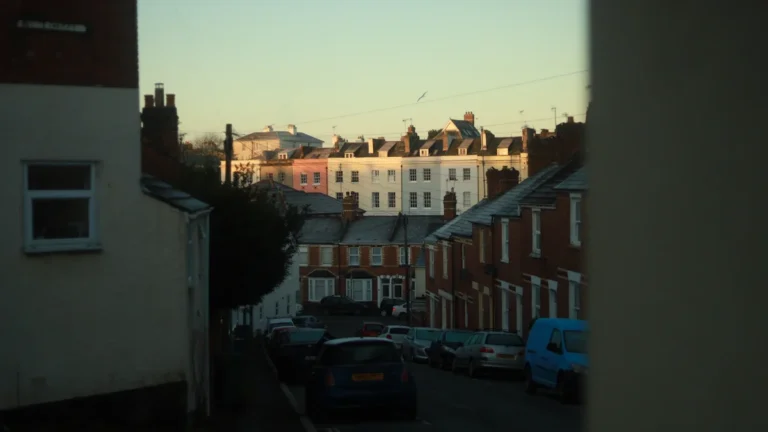 A sloped residential street in the UK featuring traditional brick terrace houses and parked cars.
