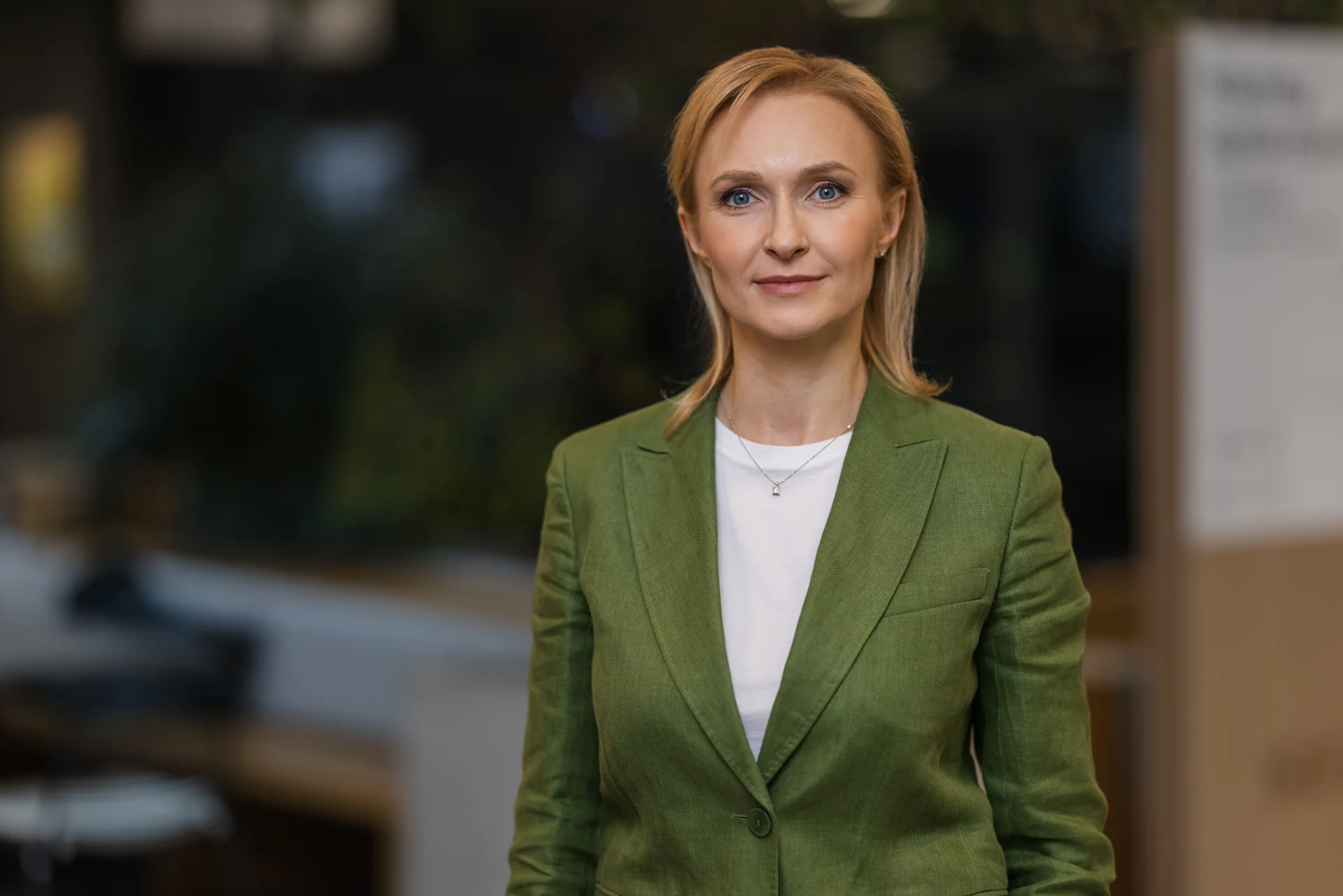 A professional blonde woman wearing a green blazer and white shirt in a blurred office environment.