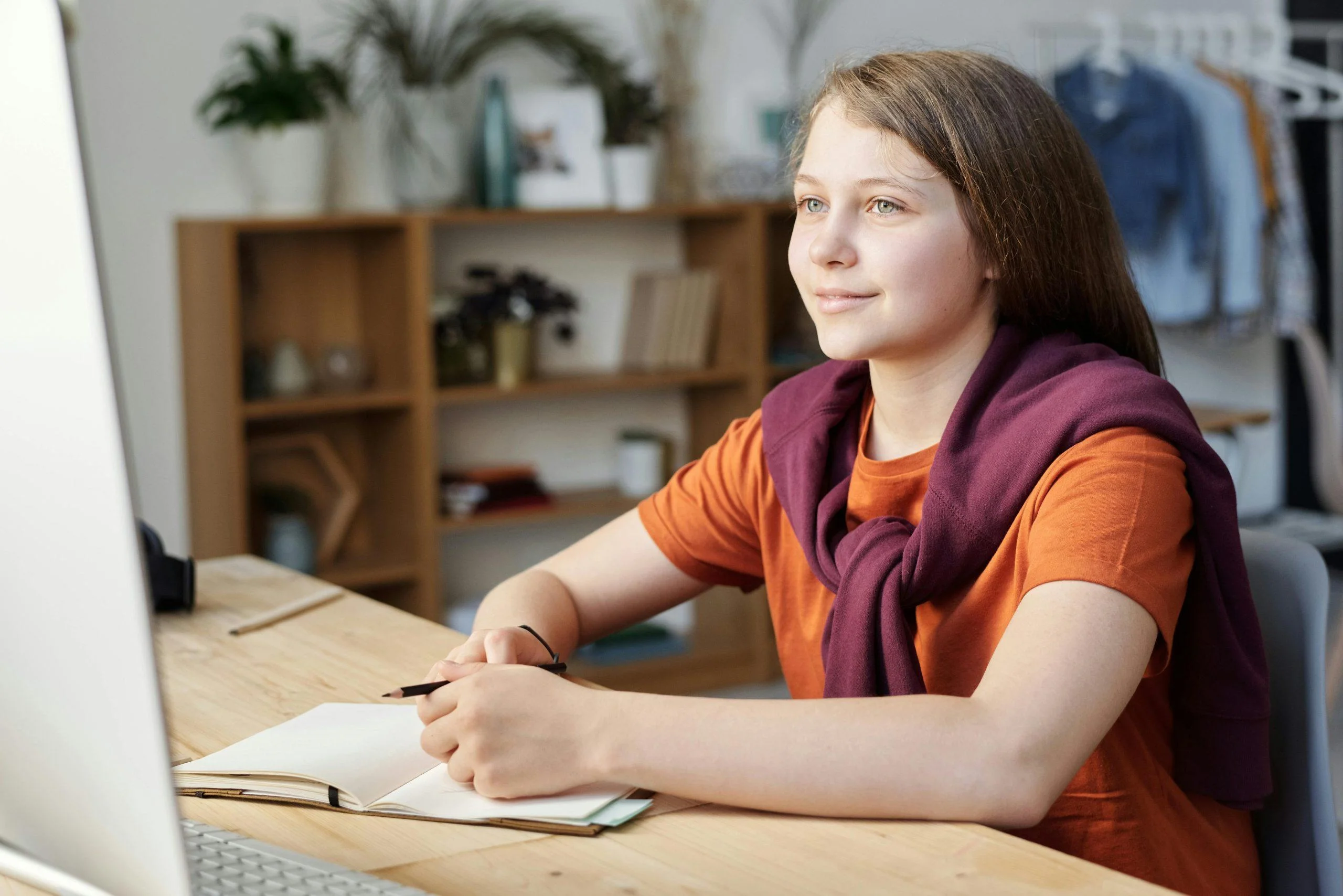A young student looks at a computer screen while holding a pencil over a notebook.