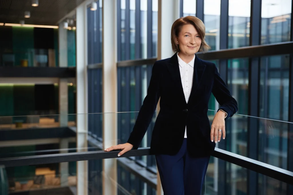 A professional woman in a navy velvet blazer stands smiling in a modern office atrium.