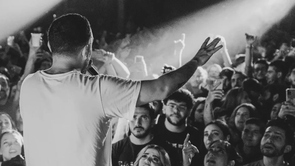 A dramatic black and white photograph from behind a lead singer performing on stage, facing a large, enthusiastic crowd illuminated by spotlights.
