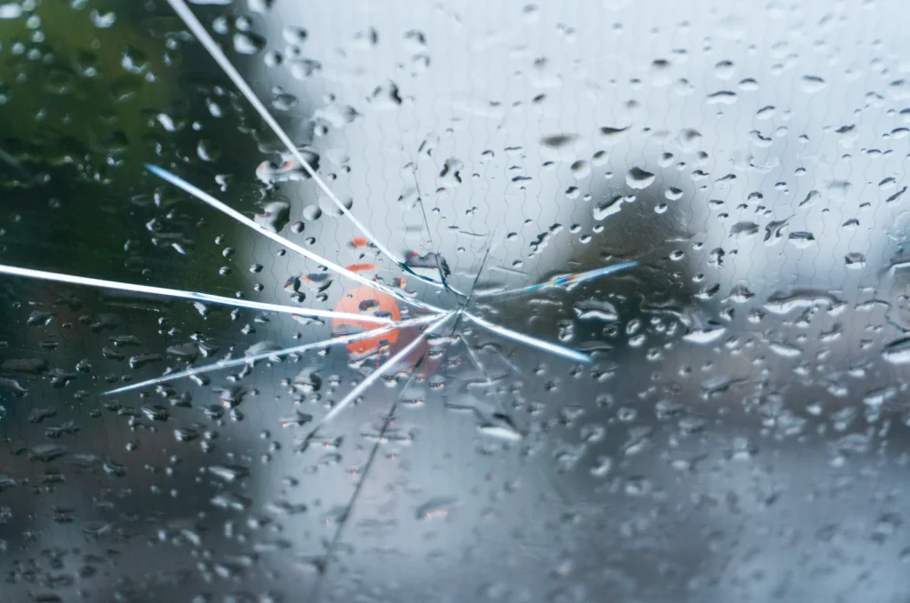 Close-up of a cracked windshield covered in raindrops with visible cracks radiating from an impact point.