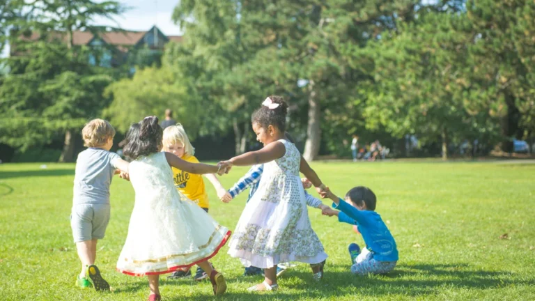Children holding hands and playing in a circle on a lush green lawn in a park.