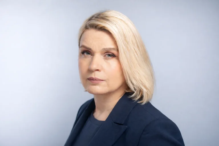 A professional blonde woman with a serious expression wearing a navy blazer against a neutral background.