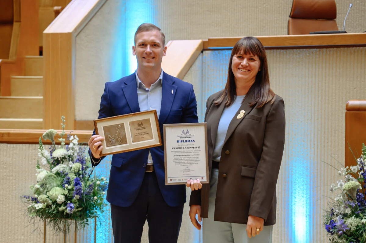 A man and a woman smiling while holding a framed award and a certificate indoors.