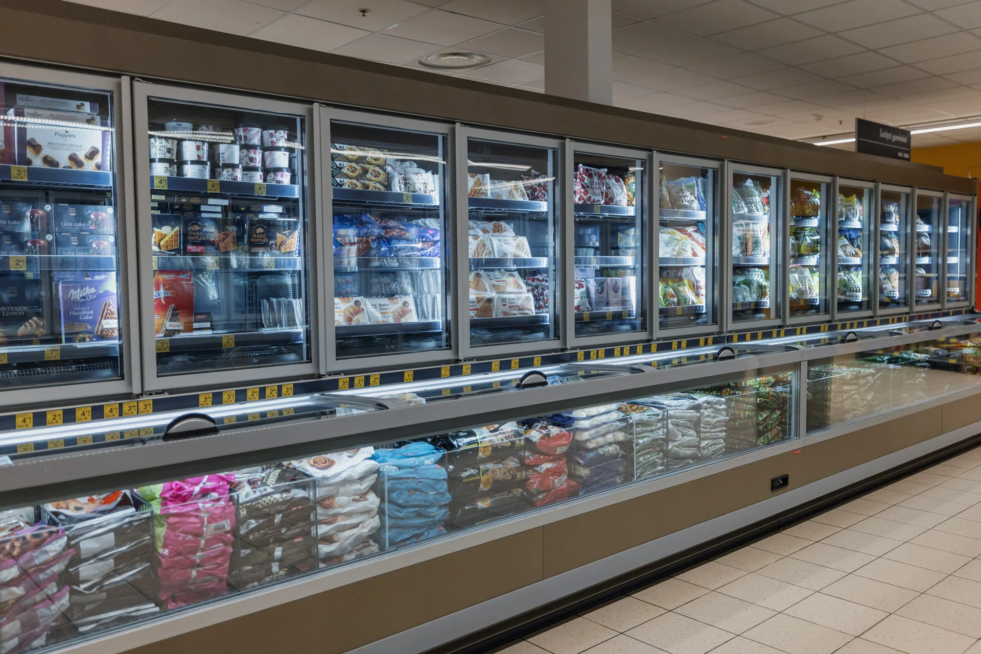 A long row of glass-door freezers in a supermarket stocked with various packaged frozen food products.