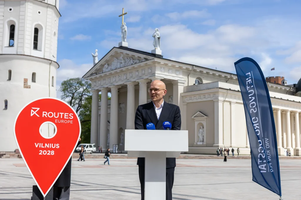 Man speaking at a podium before Vilnius Cathedral with a large red event pin.