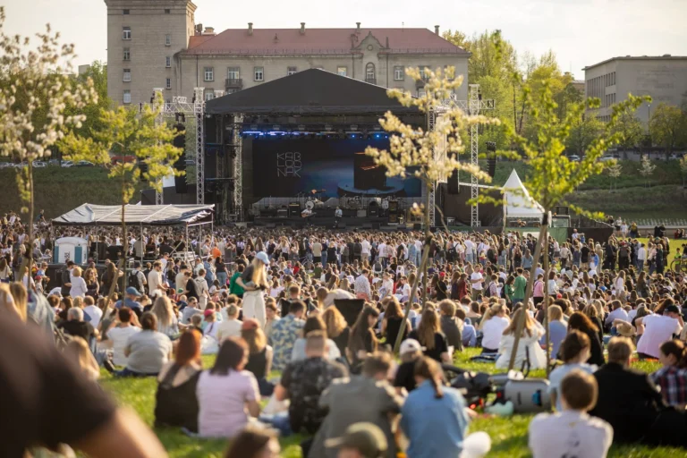 A large crowd sits on grass facing a musical performance on an outdoor city stage.