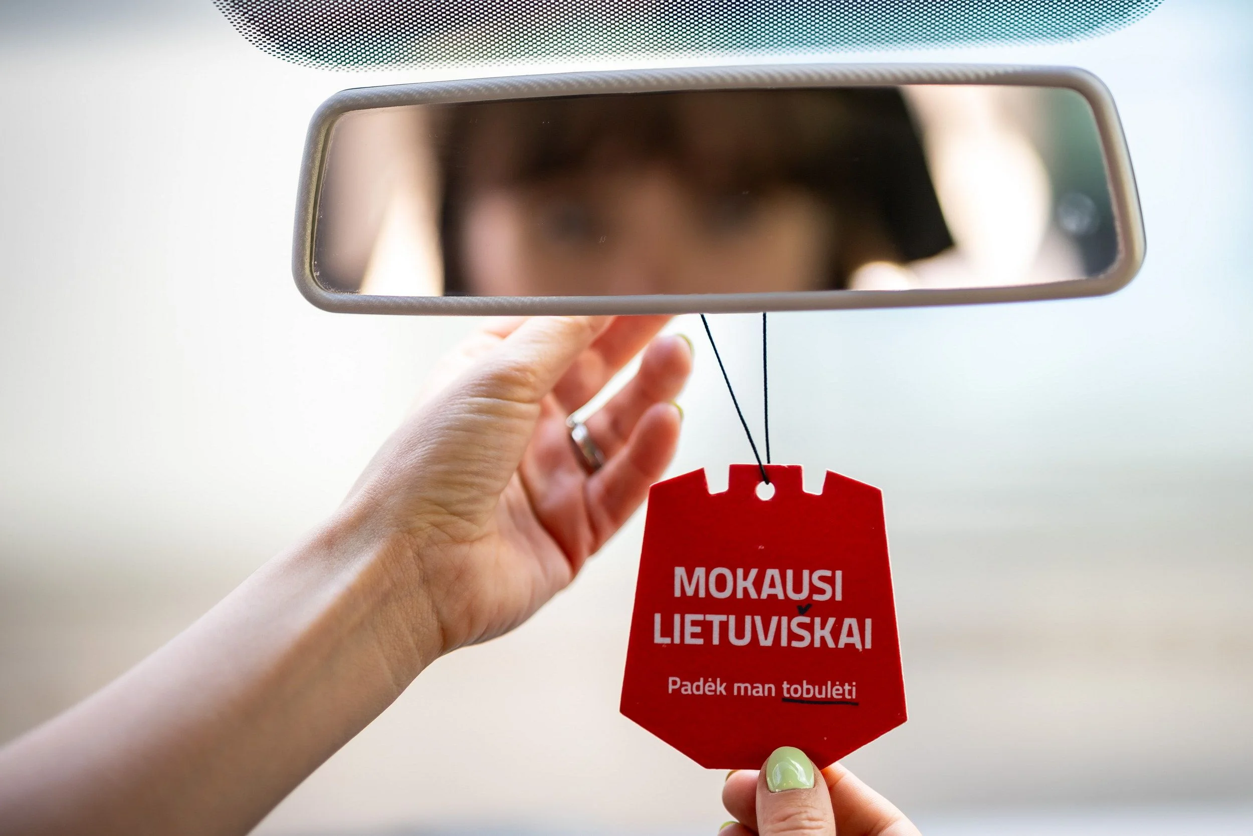 A person's hand adjusts a car rearview mirror with a red Lithuanian language learning tag.