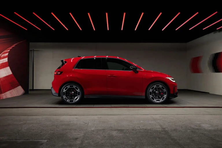 Side profile of a red compact hatchback in a dark studio with red linear ceiling lights.