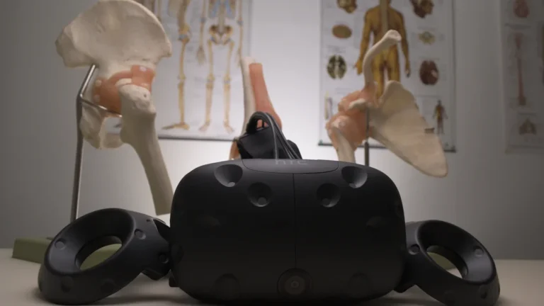 A black virtual reality headset and controllers sit on a desk in front of anatomical skeletal models.