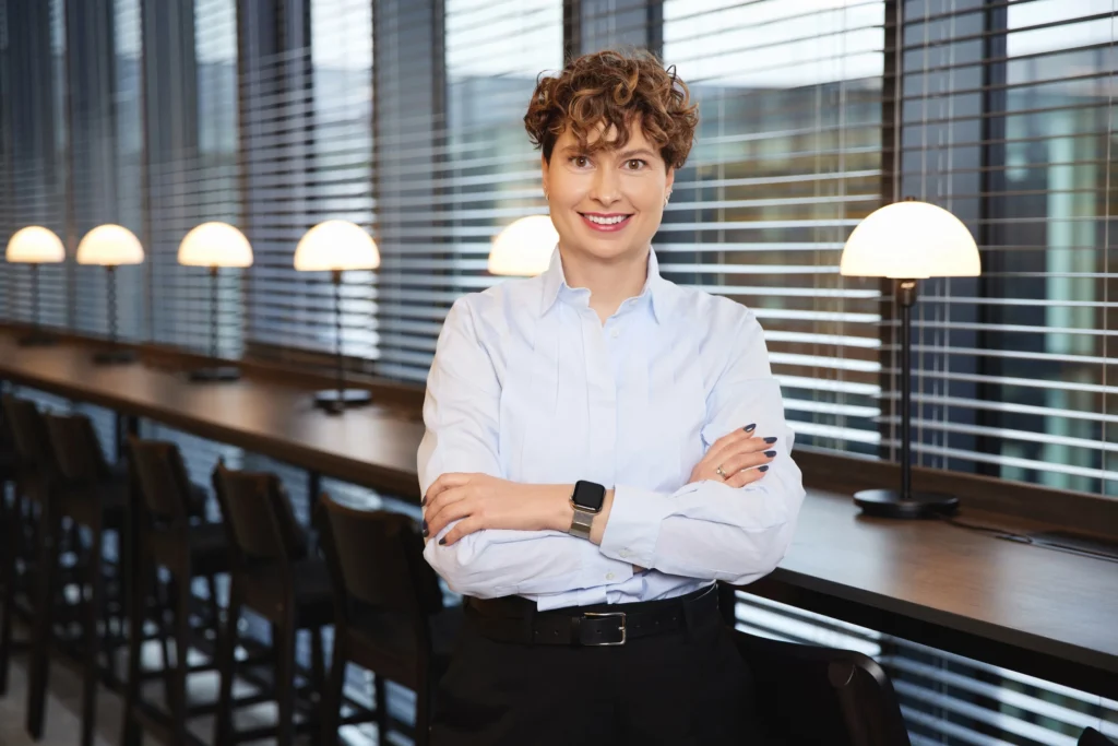 A smiling woman with short curly hair standing with crossed arms in a modern office.