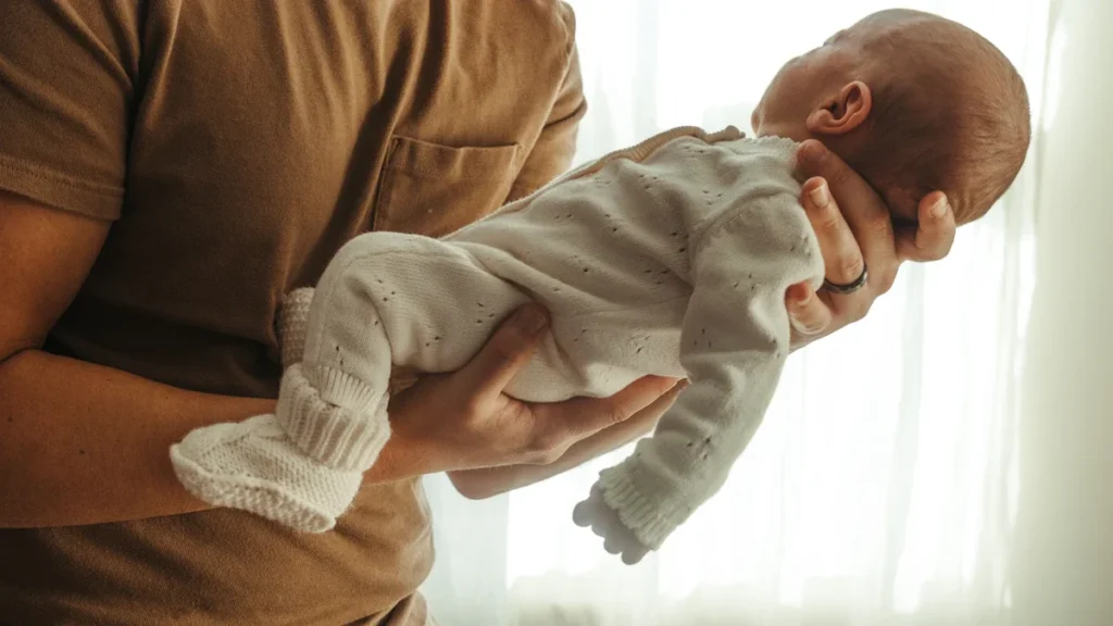 A father in a brown t-shirt gently cradling a newborn baby in a white knitted outfit.