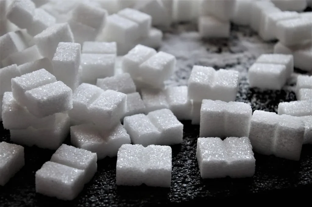 A close-up pile of white granulated sugar cubes scattered on a dark, textured surface.