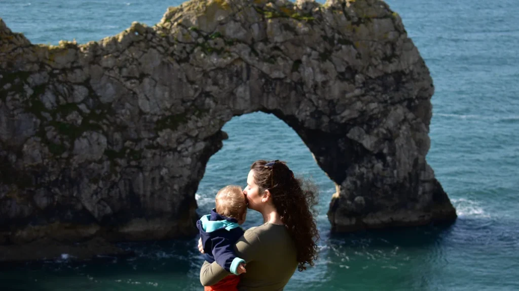A mother holds her young child while looking out at the iconic Durdle Door coastline in England.