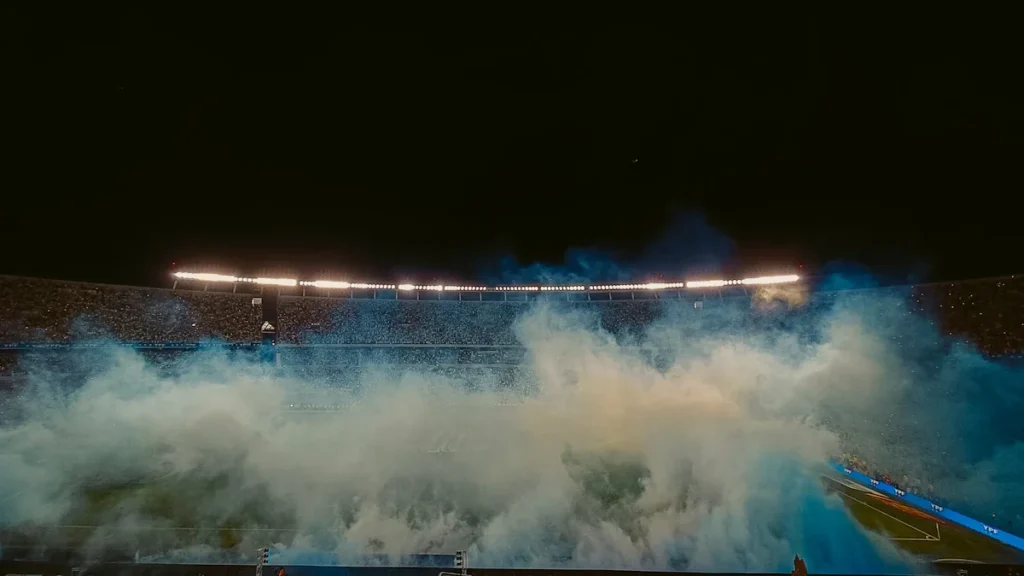 A football stadium illuminated at night with thick blue smoke rising from the field.