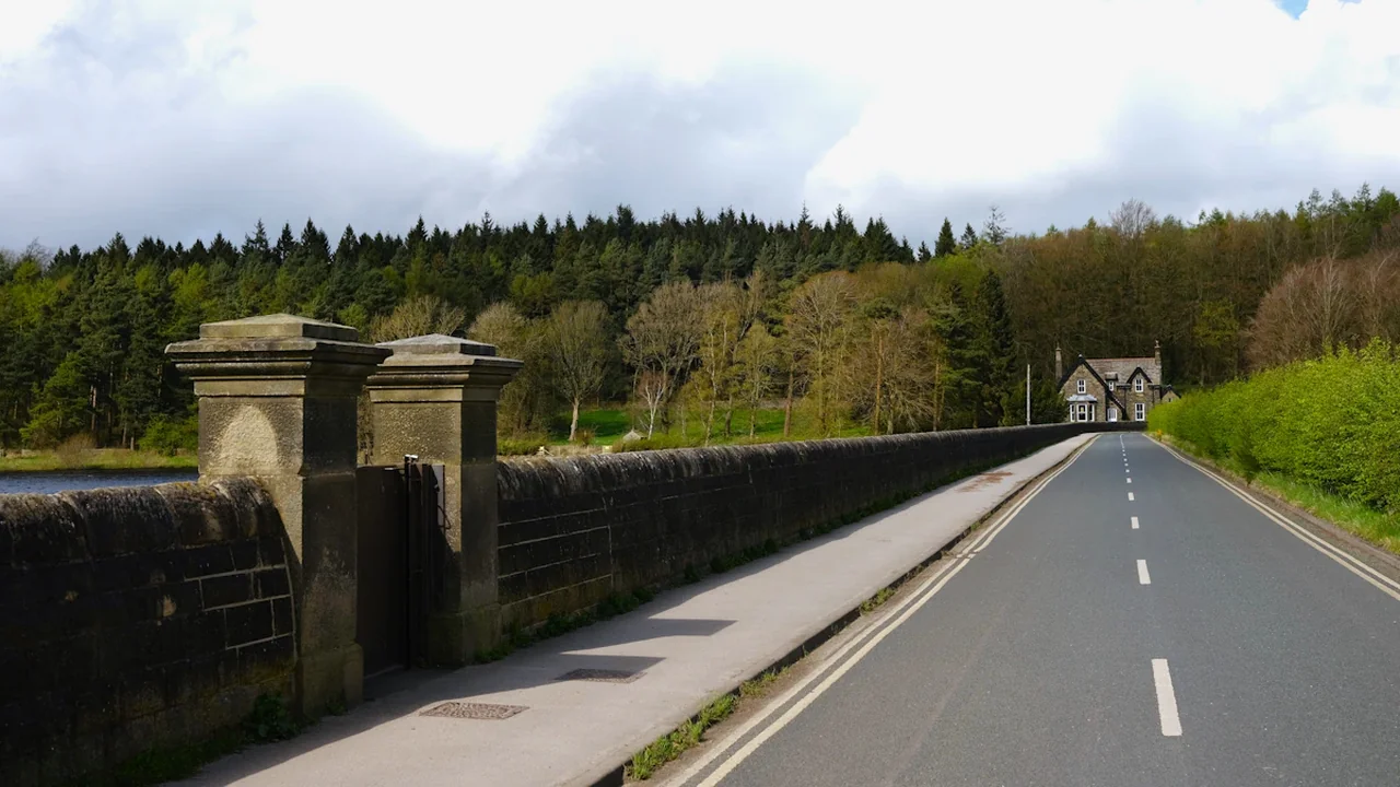 A rural UK road with a stone wall and bridge over a reservoir.
