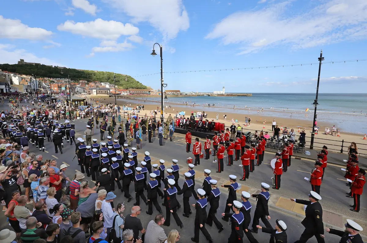 Military parade with marching personnel in uniform and red-coated musicians along a seaside promenade.