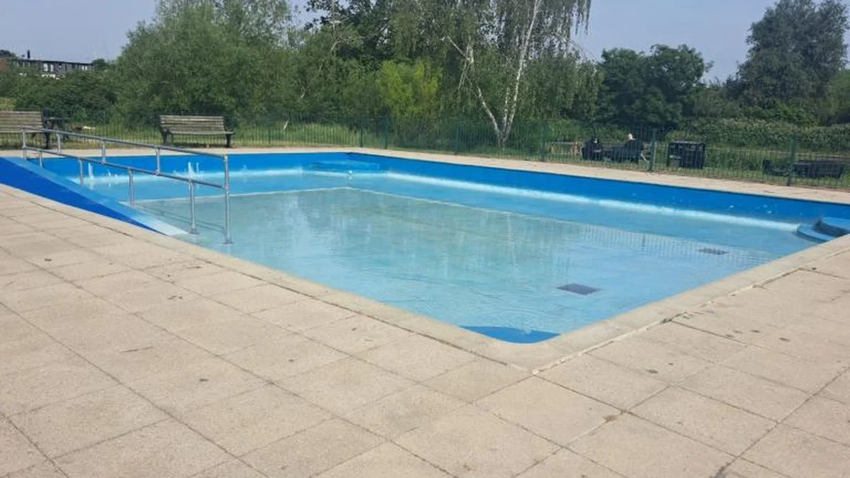 An empty outdoor swimming pool with a ramp and handrail surrounded by green trees.