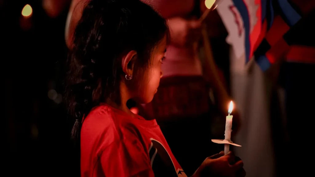 A young girl in a red shirt holds a lit candle during a night vigil.