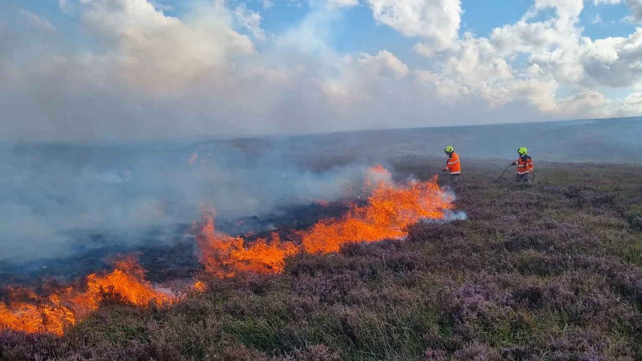 Two firefighters in bright orange gear spray water on a moorland brush fire.