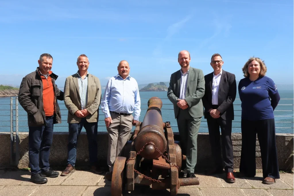 Group of six people standing outdoors by a historic cannon overlooking the ocean.