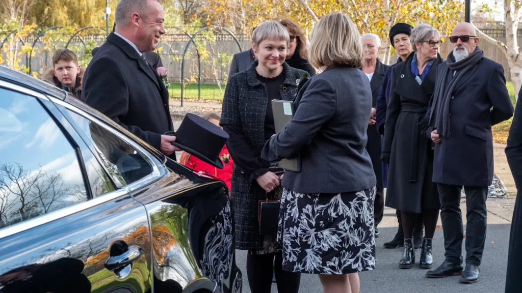 People in formal attire standing outside near a hearse in the United Kingdom.